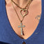 Person wearing a gold necklace with a heart pendant and a cross pendant, against a blurred outdoor background.