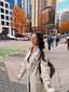 Woman in a trench coat walking on a city street with tall buildings and autumn trees.