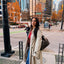 women AV Jewels founder in a trench coat standing on a city street with tall buildings and autumn trees in the background